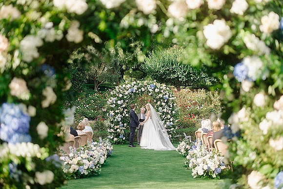Wedding ceremony with bride and groom under a floral arch, bride’s long veil flowing, framed by white roses and blue hydrangeas on a garden lawn