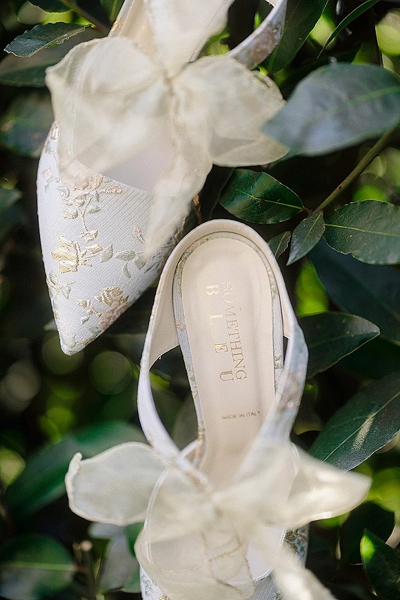 Wedding shoes with bridal heels styling, featuring metallic floral embroidery, sheer organza bows and ankle straps on green foliage backdrop