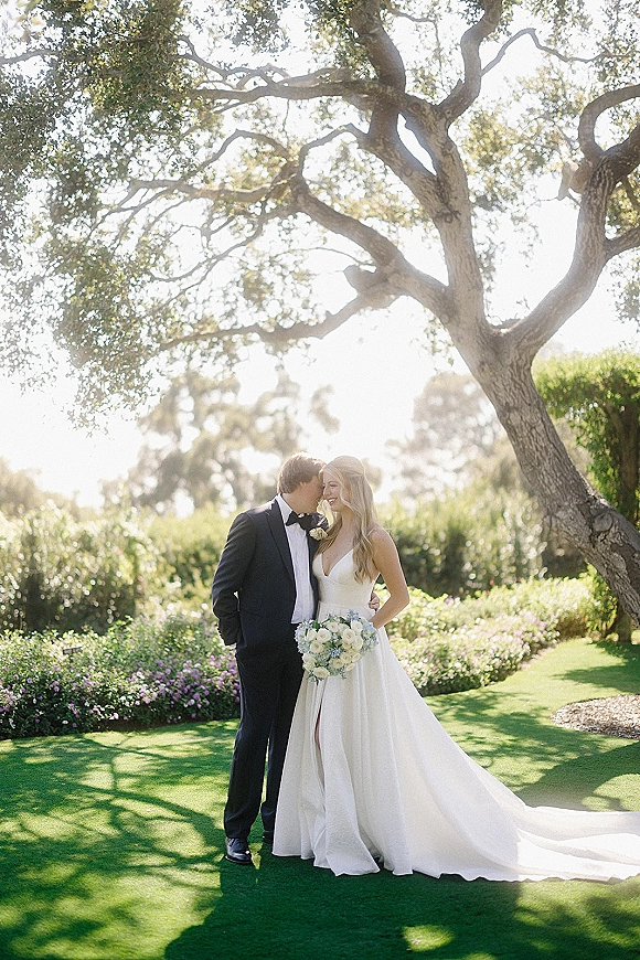 Couple portrait of bride and groom embracing with a forehead kiss, her long-train dress and bouquet under a sunlit tree on a garden lawn