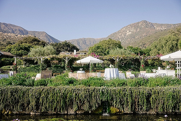 Outdoor reception setup with white linen cocktail tables and patio umbrellas, lounge seating, and florals by a pond with mountains beyond