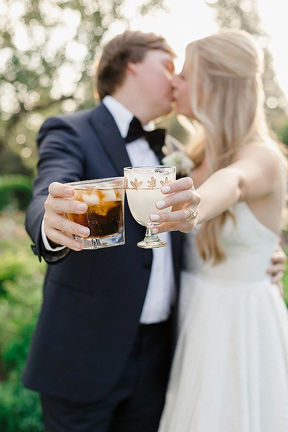 Wedding kiss portrait of bride and groom toasting with champagne coupe and whiskey glass, sunlit greenery behind, engagement ring visible