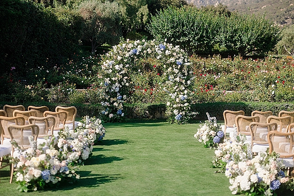 Ceremony setup for an outdoor wedding ceremony with a hydrangea and white rose floral arch, cane chairs, and flower-lined aisle on a garden lawn