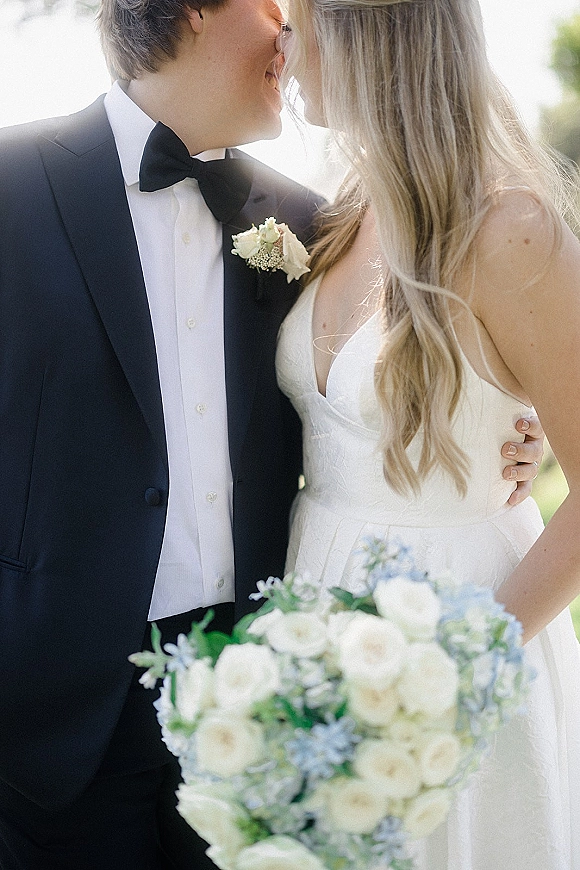 Wedding couple portrait of bride and groom close up sharing a kiss, groom in black tuxedo, bride holding white and blue bouquet in sunlit greenery