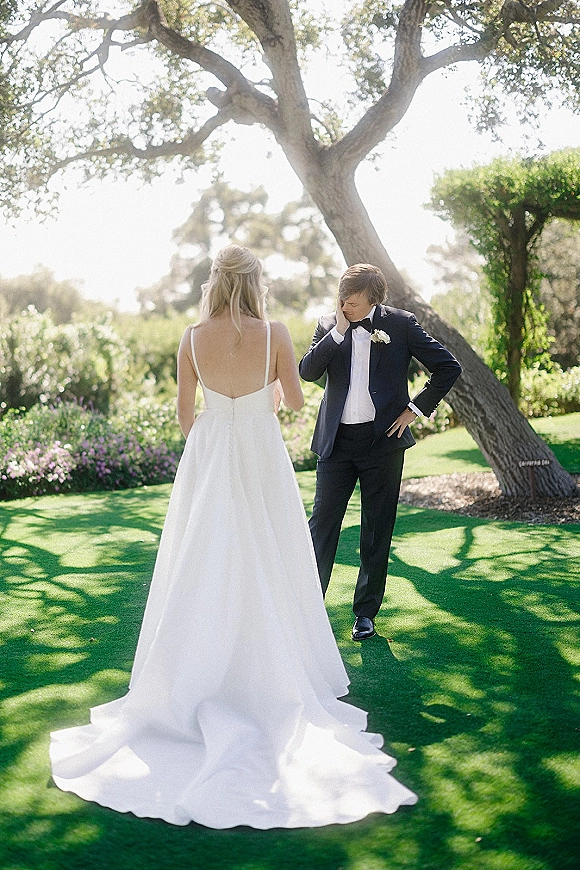 First look moment outdoors as bride in a backless wedding dress approaches groom in tuxedo wiping tears on a sunlit garden lawn under a tree