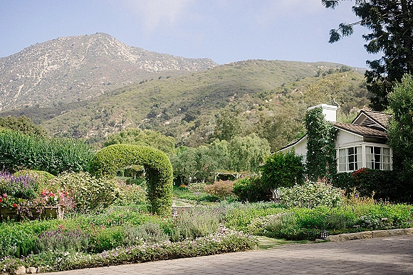 Garden wedding venue with manicured hedges framing a vine-covered arch, stone-edged paths and lights leading toward mountains and a cottage