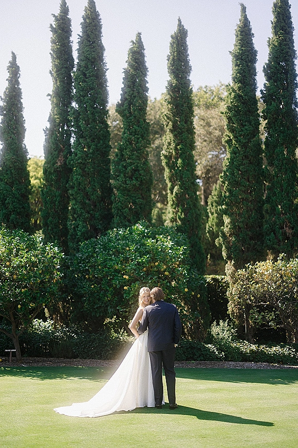 Couple portrait of bride and groom embracing from behind, groom kissing her cheek as her gown train drapes over a garden lawn.