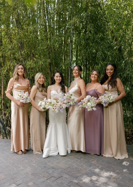 Bridesmaid group photo with bride and bridesmaids in mixed neutral satin dresses holding pastel bouquets on a stone path in bamboo grove