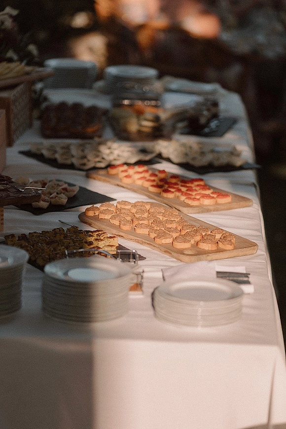 Wedding dessert table with assorted bite-size desserts on wooden serving boards over a white tablecloth, set outdoors with greenery behind
