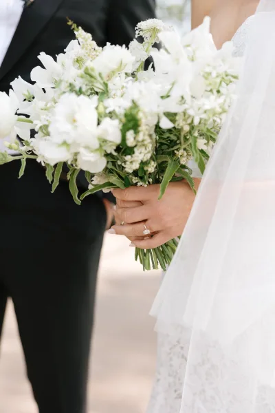 Bridal bouquet of white flowers and greenery held by a bride in veil and dress, engagement ring visible, on an outdoor walkway in soft light