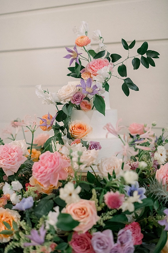 Wedding cake with tiered wedding cake layers in smooth white buttercream, accented by roses, ranunculus, clematis, and greenery on a stand