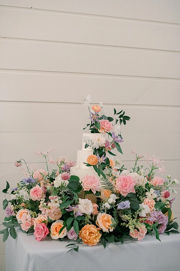 Wedding cake display featuring a tiered white wedding cake with fresh floral cake topper and greenery on a white tablecloth by a shiplap wall