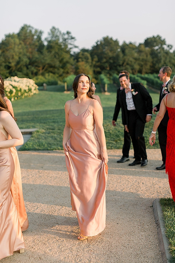Bridesmaid portrait in a blush satin bridesmaid dress with spaghetti straps, walking along a gravel path on a garden lawn at dusk