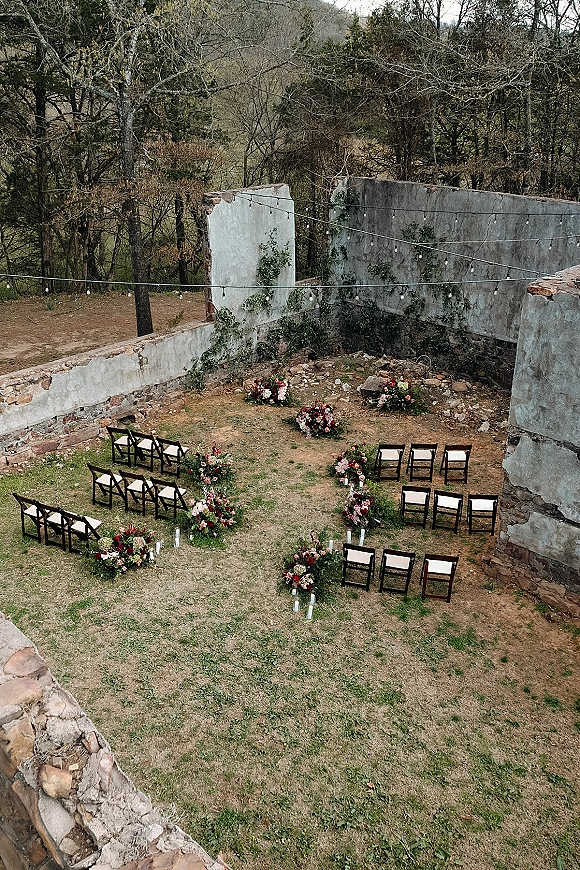 Outdoor ceremony setup in stone ruins wedding ceremony with wood folding chairs, candle-lined aisle, florals, and string lights amid trees