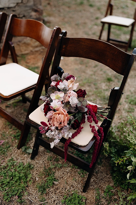 Bridal bouquet in burgundy wedding bouquet tones with roses, hellebores, orchids, and eucalyptus resting on a dark wood folding chair on grass