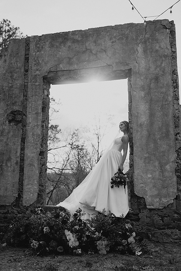 Bridal portrait of a bride in veil holding a dark floral bouquet in a stone doorway of ruins, surrounded by string lights and ground blooms
