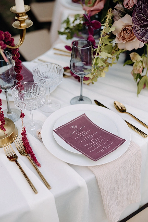 Reception tablescape with a wedding place setting, gold flatware, coupe and wine glasses, brass candlestick, and burgundy florals on white cloth
