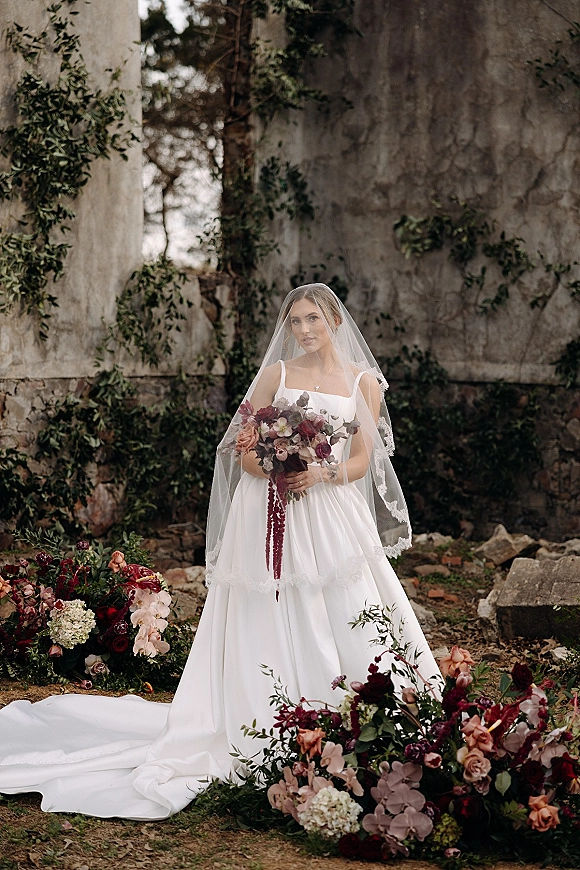 Bridal portrait of a bride holding bouquet in a white A-line dress with cathedral veil over her face, set among ivy-covered stone ruins