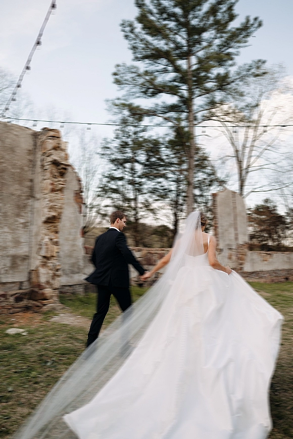 Wedding couple portrait of bride and groom walking away hand in hand, cathedral veil flowing by stone ruins with string lights at twilight