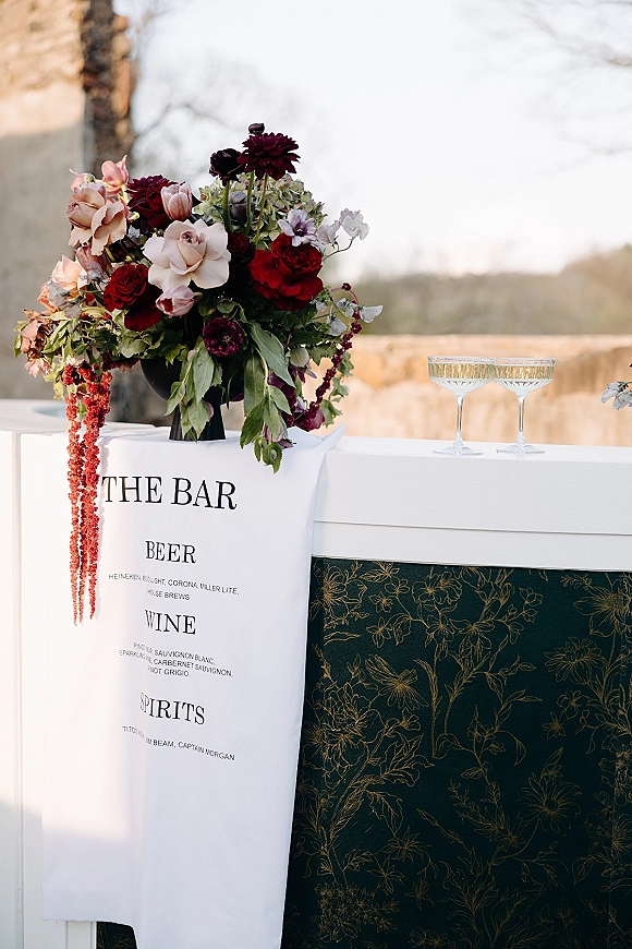 Wedding bar setup with wedding bar menu sign beside coupe glasses and dark red floral arrangement on a patterned counter against a stone wall outdoors