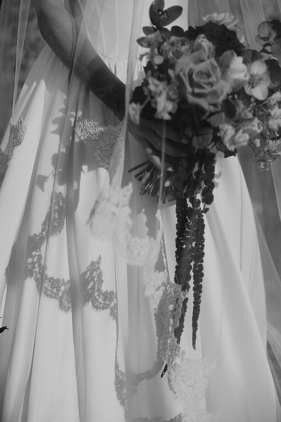 Bridal bouquet close up of roses and mixed flowers with greenery, framed by bridal veil and lace wedding dress in window light shadows