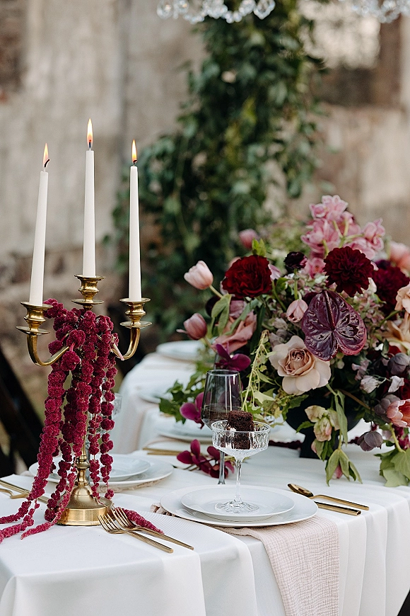 Reception tablescape with a wedding table centerpiece, brass candelabra, and white taper candles on white linen, set by a stone wall backdrop