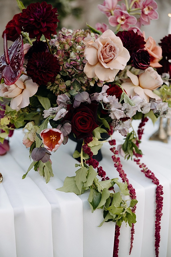 Wedding centerpiece with burgundy wedding flowers, roses, dahlias, orchids, and trailing amaranthus in a vase on a white table with blurred greenery