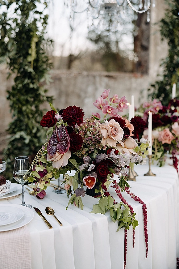 Reception tablescape with wedding floral centerpiece of burgundy blooms, blush roses, orchids, taper candles and brass candlesticks in garden setting