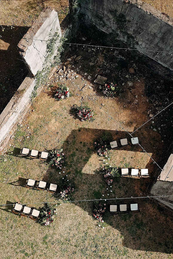 Ceremony setup with outdoor ceremony seating, white chairs lining an aisle of floral arrangements and candles under string lights in stone ruins courtyard