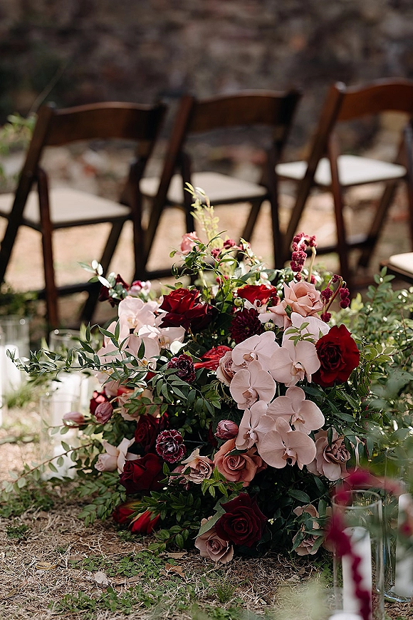 Ceremony aisle flowers with aisle ground florals featuring red and blush roses, white orchids, greenery, and candlelit glass vases by wood chairs