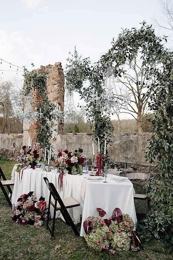 Reception tablescape with greenery arch wedding details, crystal chandeliers, taper candles and floral centerpieces set before stone wall ruins and string lights