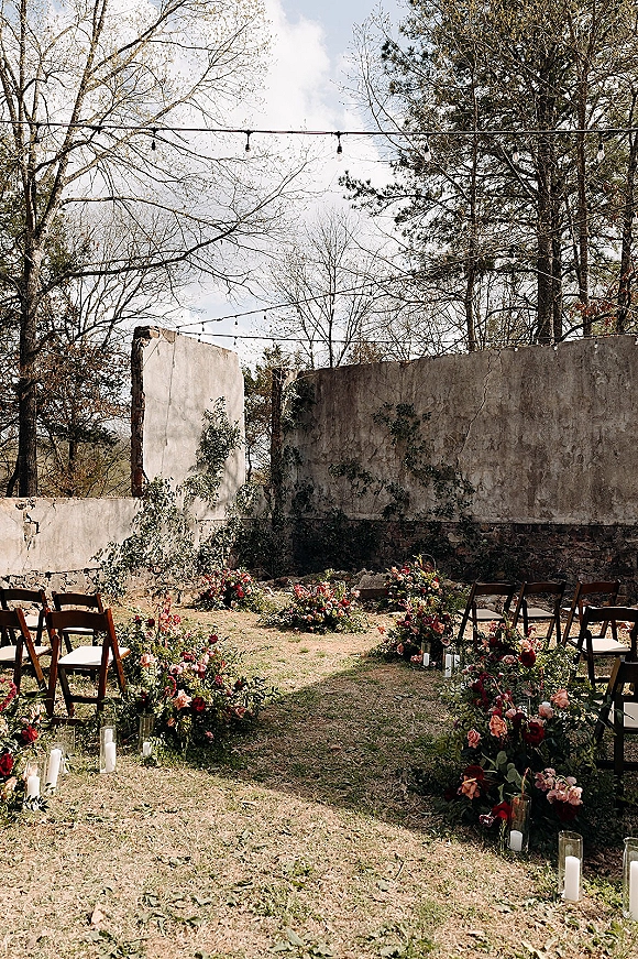 Ceremony aisle decor with a ground floral aisle of roses and greenery, pillar candles in glass cylinders, wood chairs on a lawn by ivy ruins
