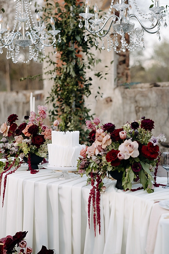 Wedding cake table with white textured cake on a stand, roses and orchids, taper candles, crystal chandeliers, and stone wall backdrop outdoors