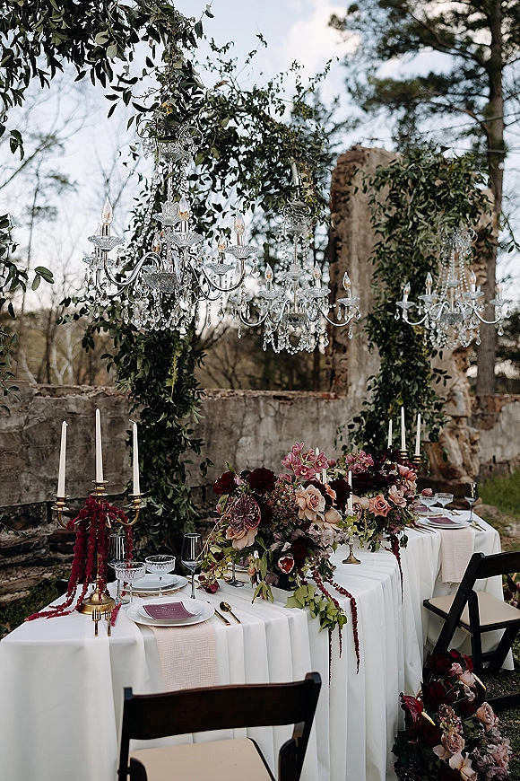 Reception tablescape with an outdoor reception table dressed in white linen, roses, taper candles, and brass candelabras beneath crystal chandeliers in a garden ruin setting