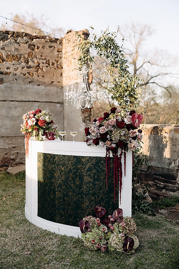Wedding bar setup with a white portable wedding bar, cascading red florals and coupe glasses under a crystal chandelier by stone ruins wall outdoors