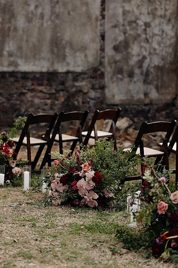 Ceremony aisle decor with aisle floral arrangements of roses and orchids, greenery, and pillar candles in glass holders beside wooden chairs on a lawn