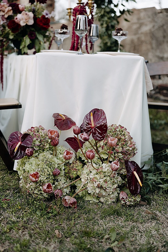 Reception sweetheart table with sweetheart table decor, white linen, hydrangea florals, brass candlesticks, and champagne coupes on a lawn by a stone wall