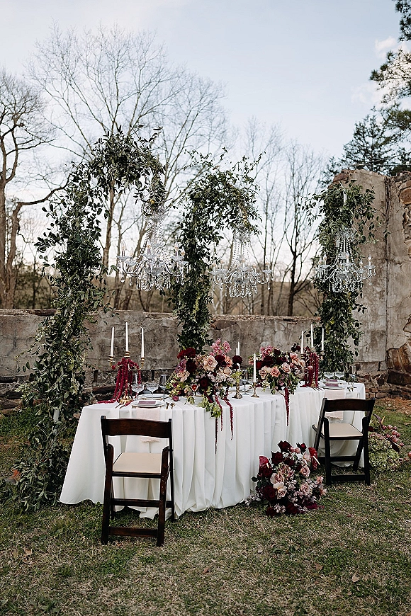 Reception tablescape with outdoor reception table styling, white linens, taper candles and floral centerpieces beneath crystal chandeliers by stone ruins