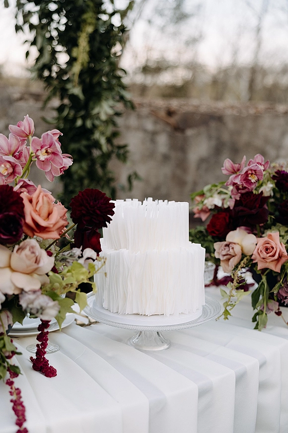 Wedding cake with textured buttercream on a glass stand, surrounded by orchid florals and tableware in an outdoor stone-wall setting