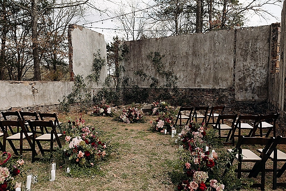 Ceremony setup with outdoor ceremony seating, black folding chairs flanking lush aisle floral arrangements and candles before a ruined stone wall