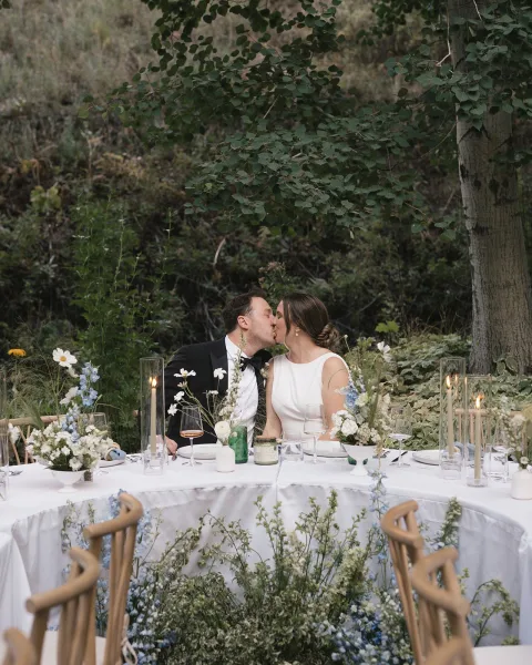 Wedding kiss as bride in a wedding dress and groom in tuxedo lean over a candlelit sweetheart table with wildflowers, greenery behind