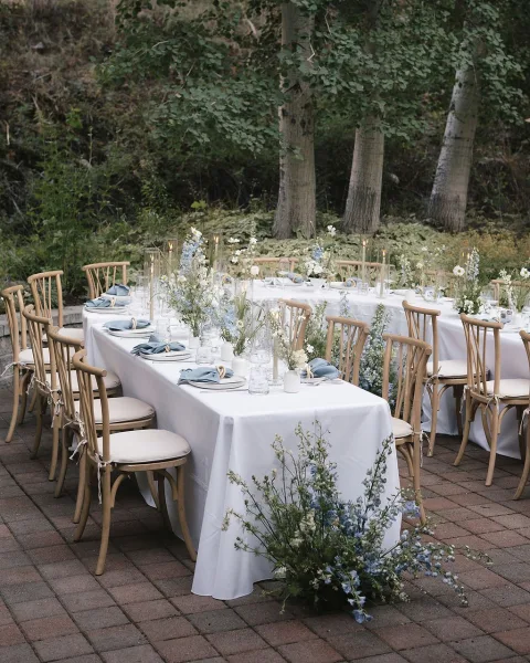 Reception tablescape with outdoor reception tables set in white linens, blue napkins, taper candles, and wildflower vases on a garden patio