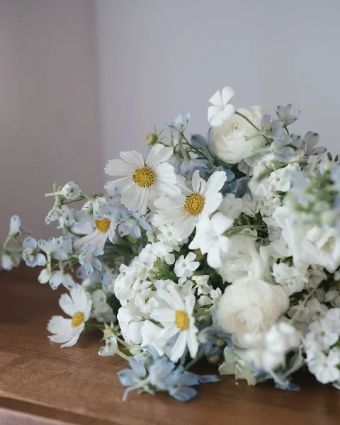 Bridal bouquet with white and blue bouquet blooms and greenery resting on a wooden tabletop against a neutral wall