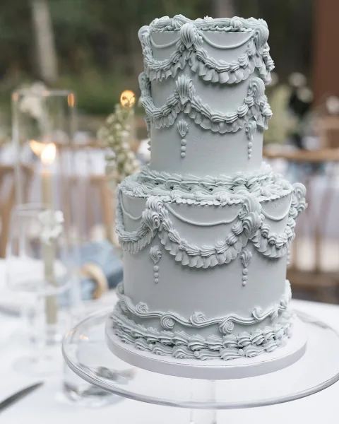 Wedding cake with tiered wedding cake ruffle frosting and piped garlands on a stand, flanked by taper candles at an outdoor reception