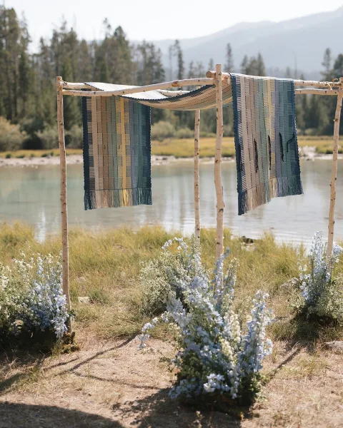 Ceremony backdrop with birch wedding arch, woven rugs and draped fabric, framed by lakeside grass, pine trees, and mountains