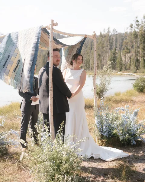Wedding ceremony moment with bride and groom holding hands under a fabric-draped arch, wildflowers beside a mountain lake shoreline