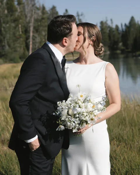 Wedding kiss portrait of bride and groom kissing, bride holding white wildflower bouquet by a lake with evergreen trees and blue sky