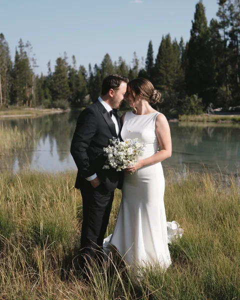 Couple portrait of bride and groom holding a wildflower bouquet, touching foreheads in tall grass by a lake and pine trees under blue sky
