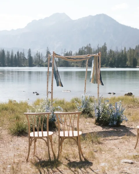Ceremony setup for a lake wedding ceremony with a wood arch draped in fabric and florals, chairs facing a mountain lake and pines