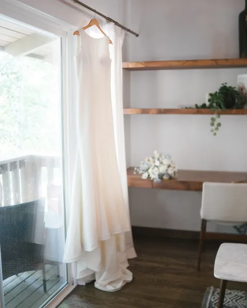 Wedding dress hanging by window in natural light, minimalist bridal gown on wooden hanger with greenery and bouquet on shelves