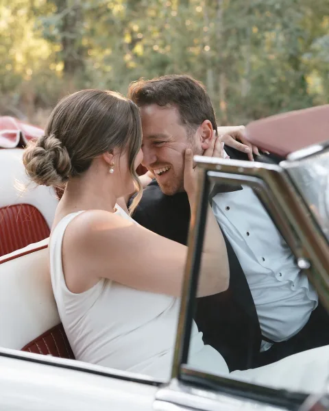 Wedding couple portrait of bride and groom in car, laughing as she holds his face in a vintage convertible under sunlit trees